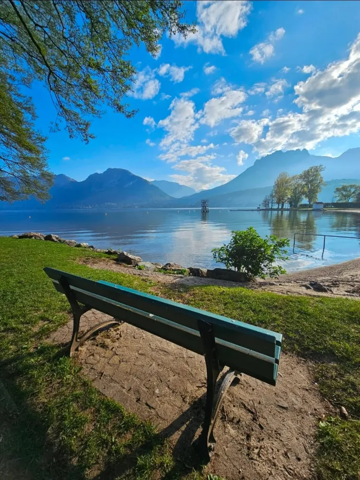 Banc vert au bord d'un lac calme, face à un plongeoir au loin et à des montagnes sous un ciel bleu nuageux.